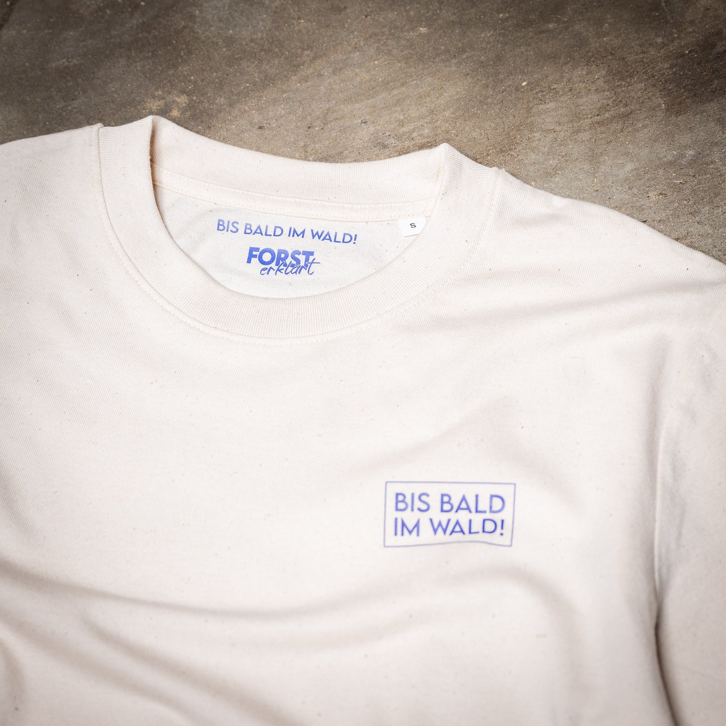 Eine Nahaufnahme des Waldshirts Leberblümchen. Man sieht die Drucke auf der Vorderseite. In Leberblümchen-Blau BIS BALD IM WALD! in einer Box auf der Brust links und im Kragen. Im Kragen ist zudem das Forst erklärt-Logo.